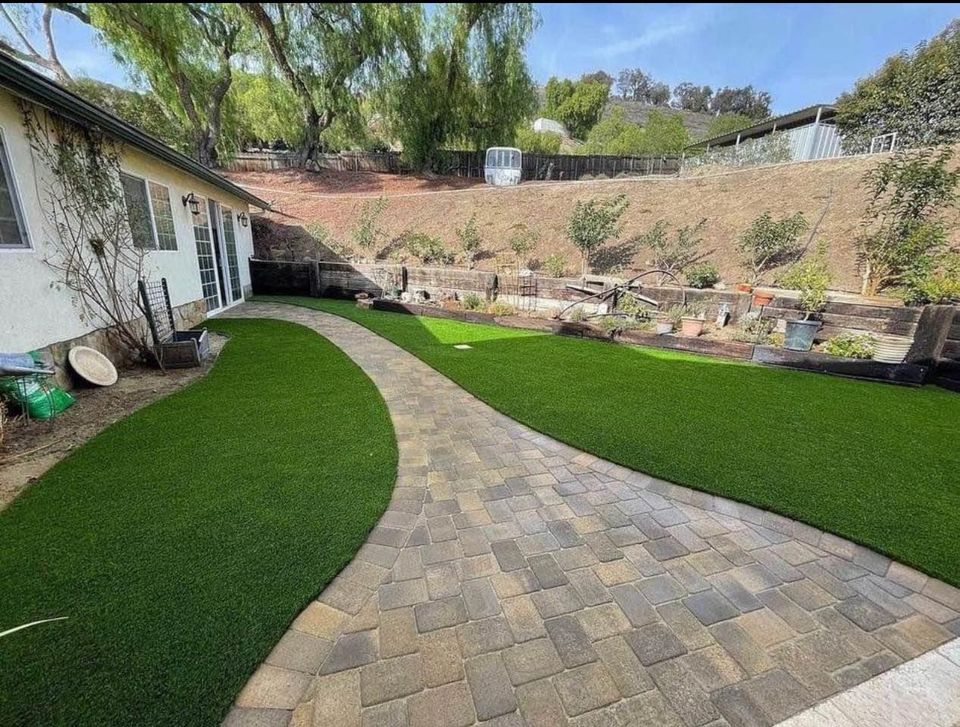 A backyard in Paradise Valley features a stone pathway leading to a garden. Flanking the path is low-maintenance artificial grass, offering lush greenery year-round. A variety of plants and trees thrive in the raised garden bed, with a house visible on the left.