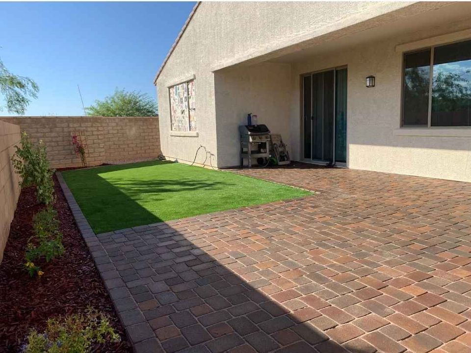 A backyard patio in Paradise Valley, AZ, features red brick paving and a small low-maintenance grassy area. A barbecue grill stands by the house with light-colored siding. A sliding glass door leads inside, while a tall brick fence encloses the yard, with garden beds bordering the patio.