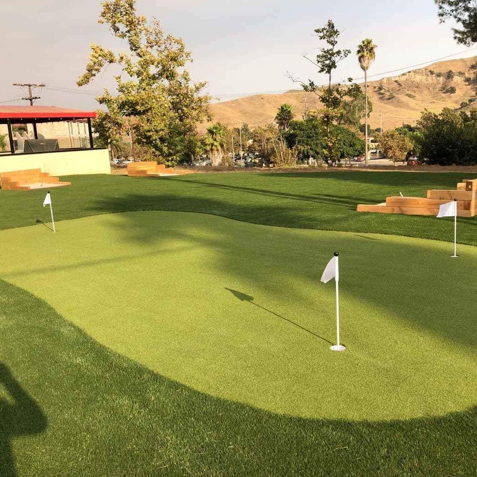 A mini-golf course with three small white flags on smooth putting greens. Surrounding the course are wooden steps, trees, and hills under a clear sky. A red-roofed building is visible in the background, perfect for any golf enthusiast seeking a scenic play area.