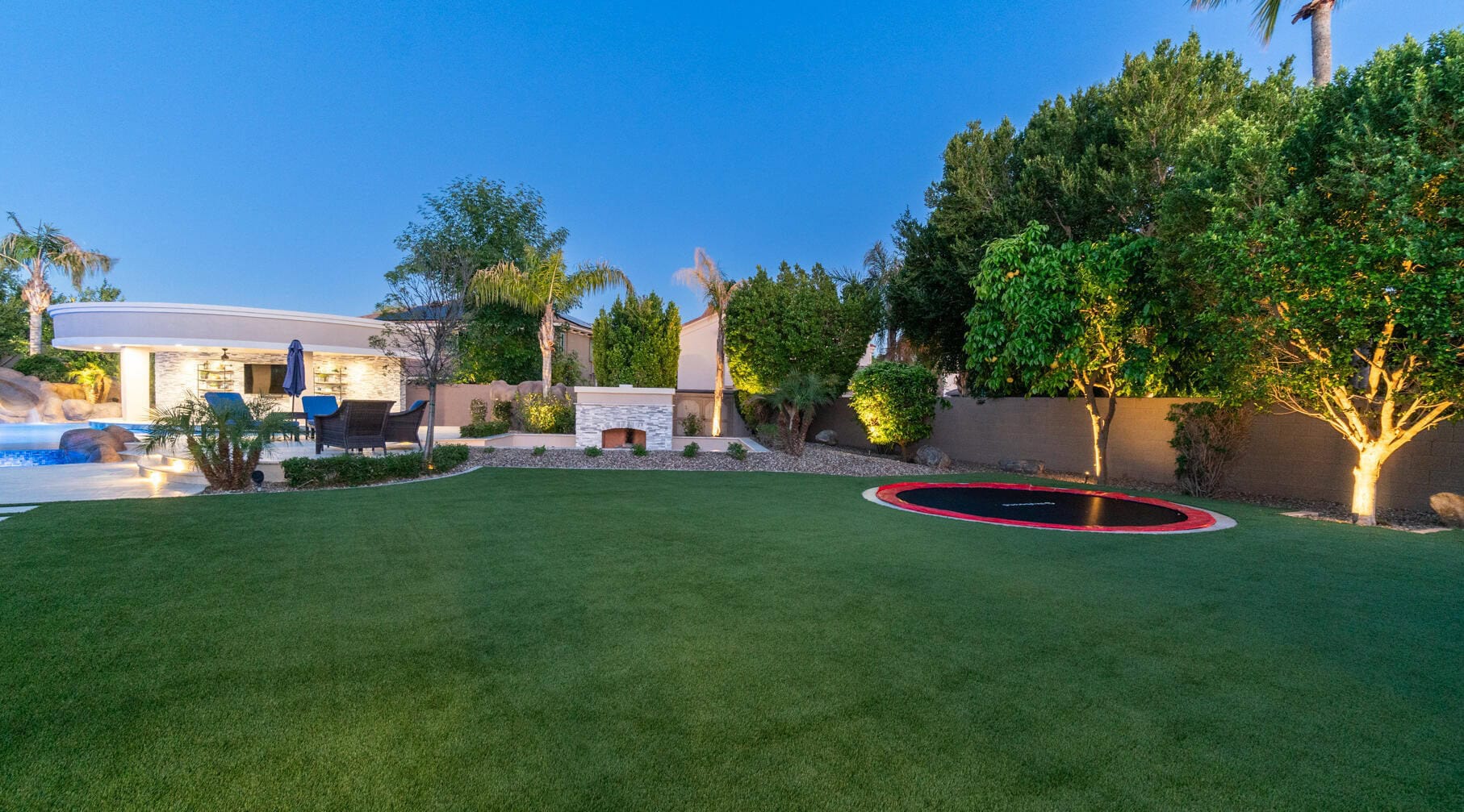 A spacious backyard featuring a well-manicured lawn of low-maintenance artificial grass with a trampoline set into the ground, surrounded by trees and bushes. A pool is visible on the left near a patio area with outdoor furniture and a white stone fireplace under Paradise Valley AZ's clear blue sky.