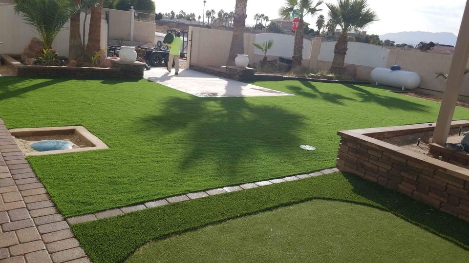 A backyard paradise with lush artificial grass and surrounded by brick planters and palm trees. A paved walkway and stone patio lead to someone working near a small fountain. In the background, Paradise Valley's desert scenery with mountains creates a stunning view.