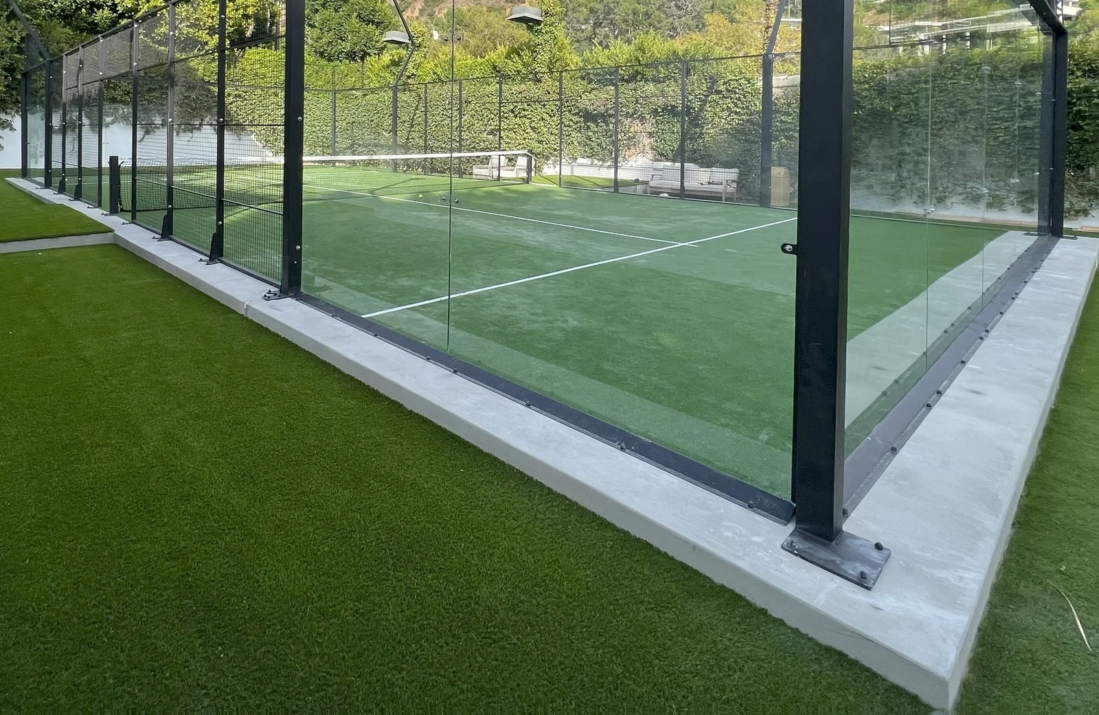 A padel court enclosed by glass walls on a sunny day, featuring cutting-edge artificial turf installations. The court is surrounded by well-maintained grass, with trees and hedges in the background, reminiscent of the pristine landscapes often found in Scottsdale AZ or Paradise Valley.