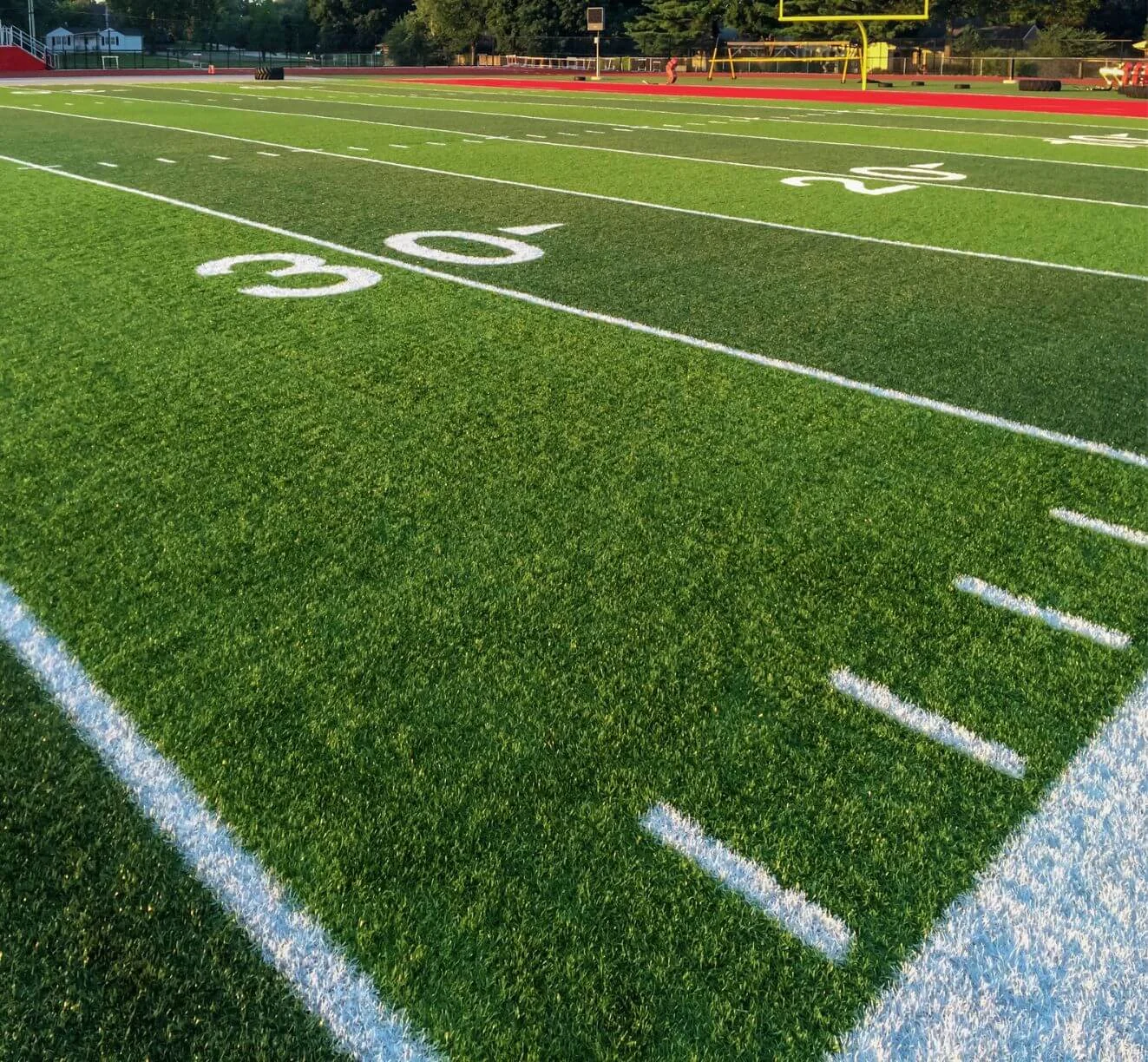 A well-maintained American football field under bright sunlight in Paradise Valley, featuring the white yard lines and numbers. The 30-yard line on the artificial turf is visible, with goalposts and a red track surrounding the field in the background.