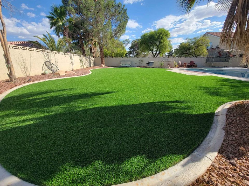 A Paradise Valley lawn boasts a lush green expanse surrounded by trees and a stone border. To the right, the sparkling swimming pool reflects the clear blue sky above. A wall lines the back edge, with plants providing shade, completing this serene paradise.