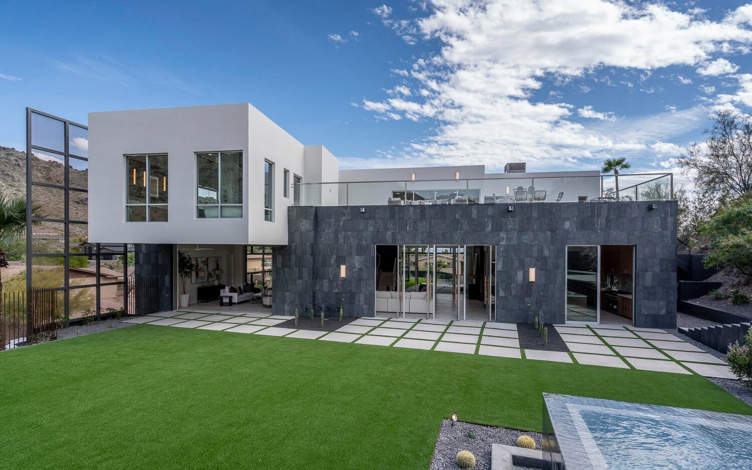 Modern two-story house with a sleek design, featuring large windows and a mix of white and dark gray exteriors. It boasts a spacious Paradise Valley lawn with seamless pet turf installation and a small reflective water feature, set against a partly cloudy sky.