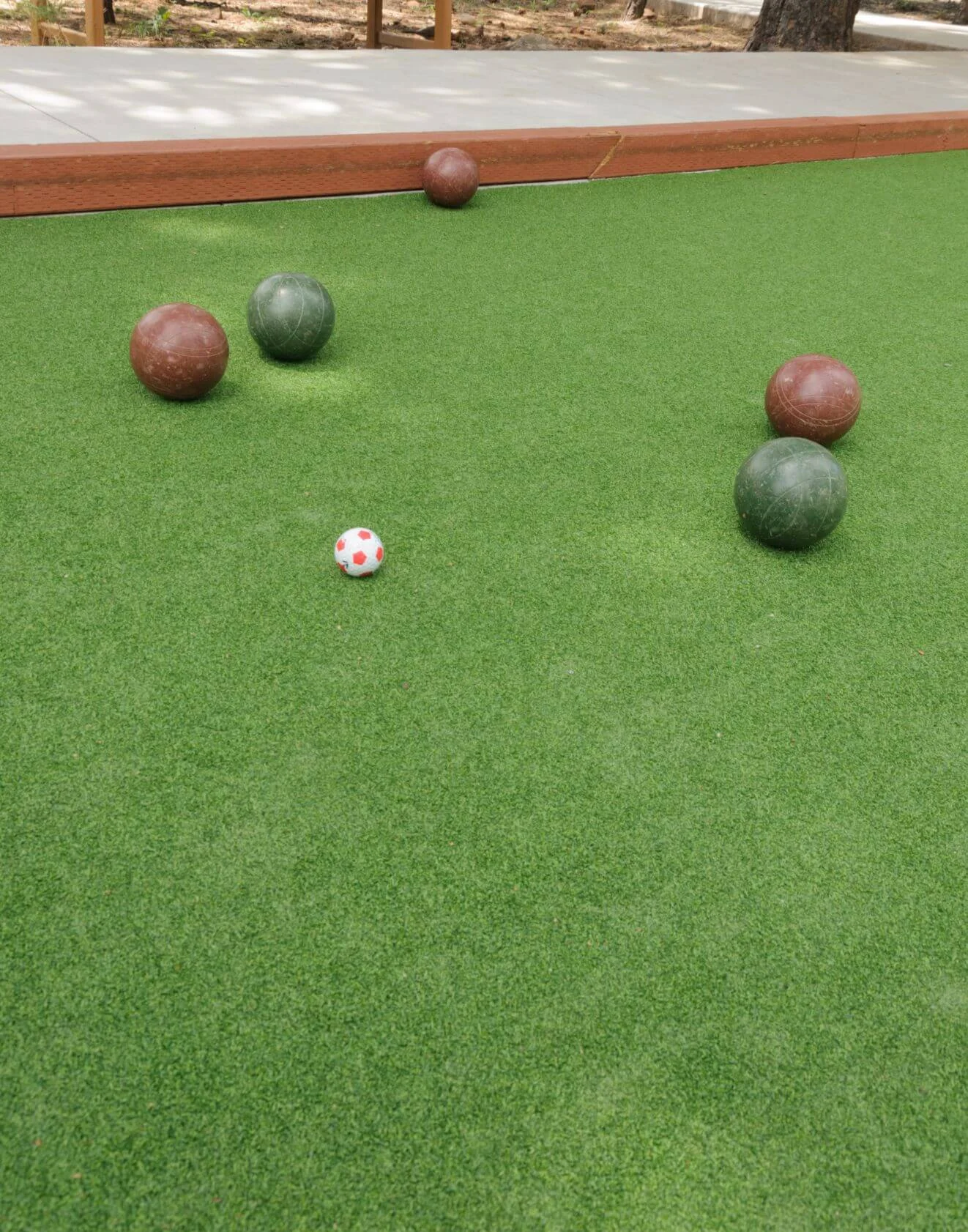 An outdoor bocce ball court in Scottsdale features three red balls, two green balls, and a small white ball with red polka dots on lush artificial turf. The court is bordered by wooden edges, with a paved area and trees in the background.