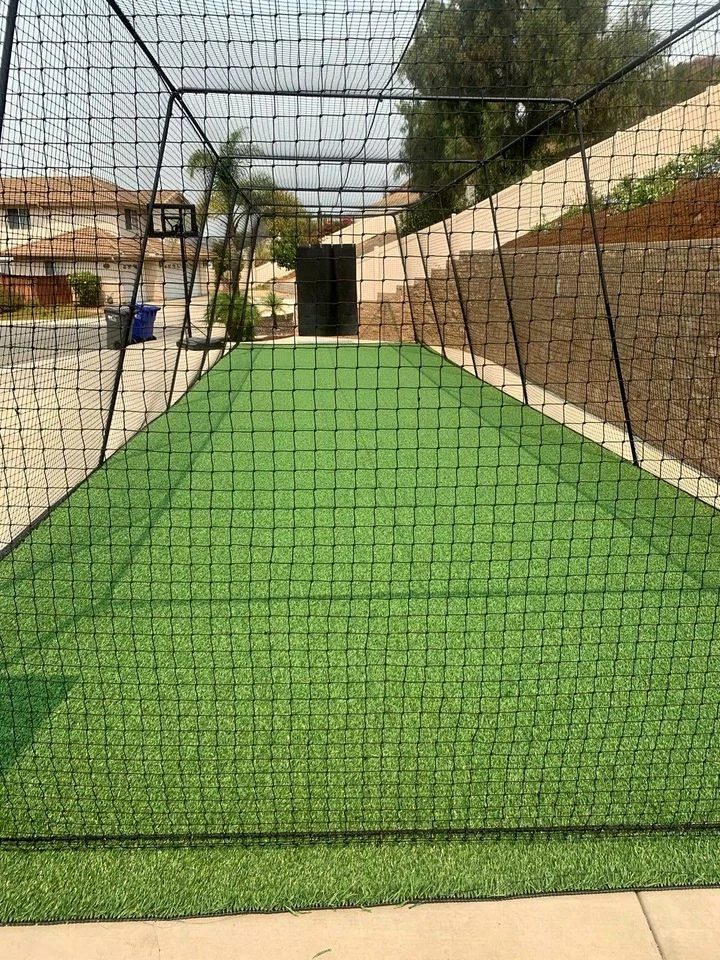 A long rectangular outdoor batting cage with artificial turf installations and surrounding netting, set near a residential area in Paradise Valley with a clear sky and trees in the background.