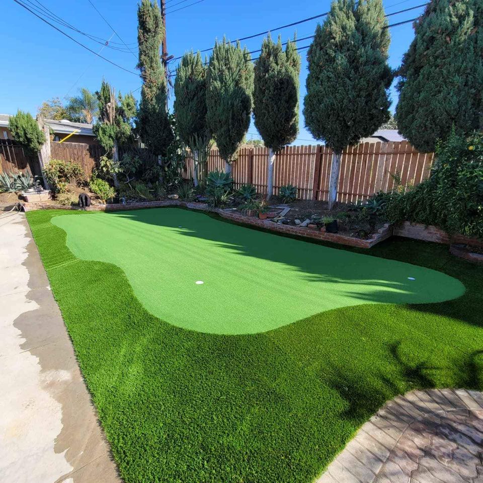 A backyard paradise for the golf enthusiast, featuring a neatly maintained synthetic putting green surrounded by lush artificial grass. Framed by tall evergreen trees and a wooden fence, the clear blue sky above enhances this serene garden setting.