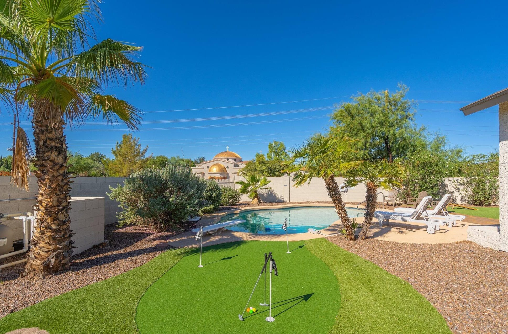 The backyard, perfect for a golf enthusiast, features putting greens with several holes, surrounded by desert landscaping. A small pool with lounge chairs is nestled beside swaying palm trees. In the background, a stucco building with a dome stands under the clear blue sky.