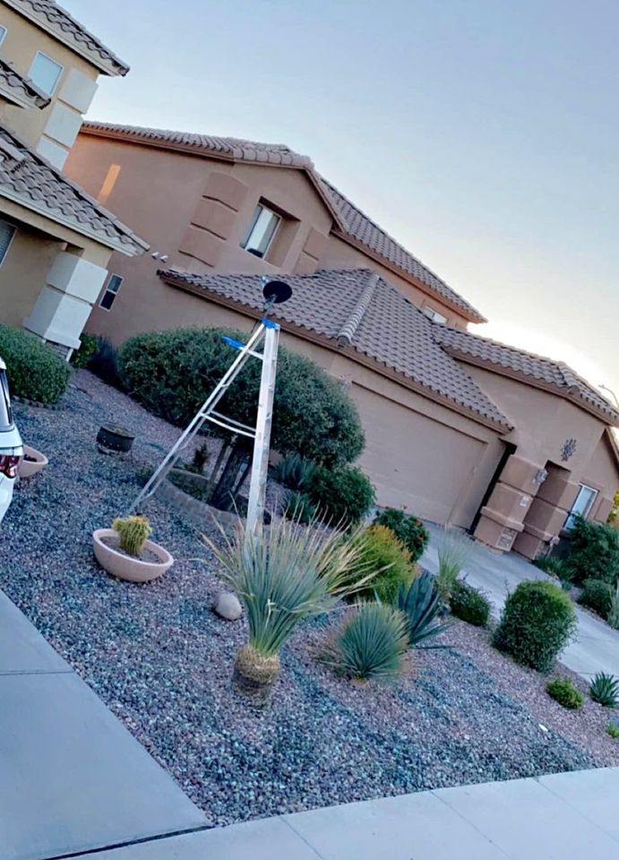 A suburban house in Paradise Valley, AZ, boasts a desert landscape garden with succulents and shrubs. A ladder with a black hat stands amid the scene. The house features beige walls, a brown tiled roof, and artfully arranged landscape installations that include artificial grass.