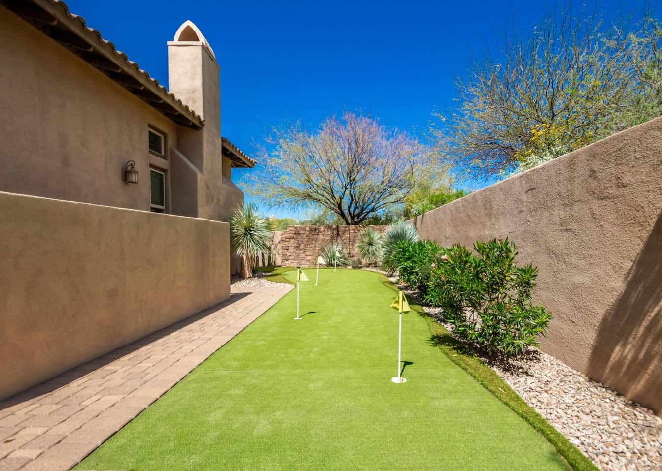 A narrow backyard ideal for a golf enthusiast, featuring a small putting green with three flag sticks made of durable artificial grass. The area is bordered by a stone path on one side and a desert landscape with shrubs and a tree on the other, all enclosed by a stucco wall under a clear blue sky.