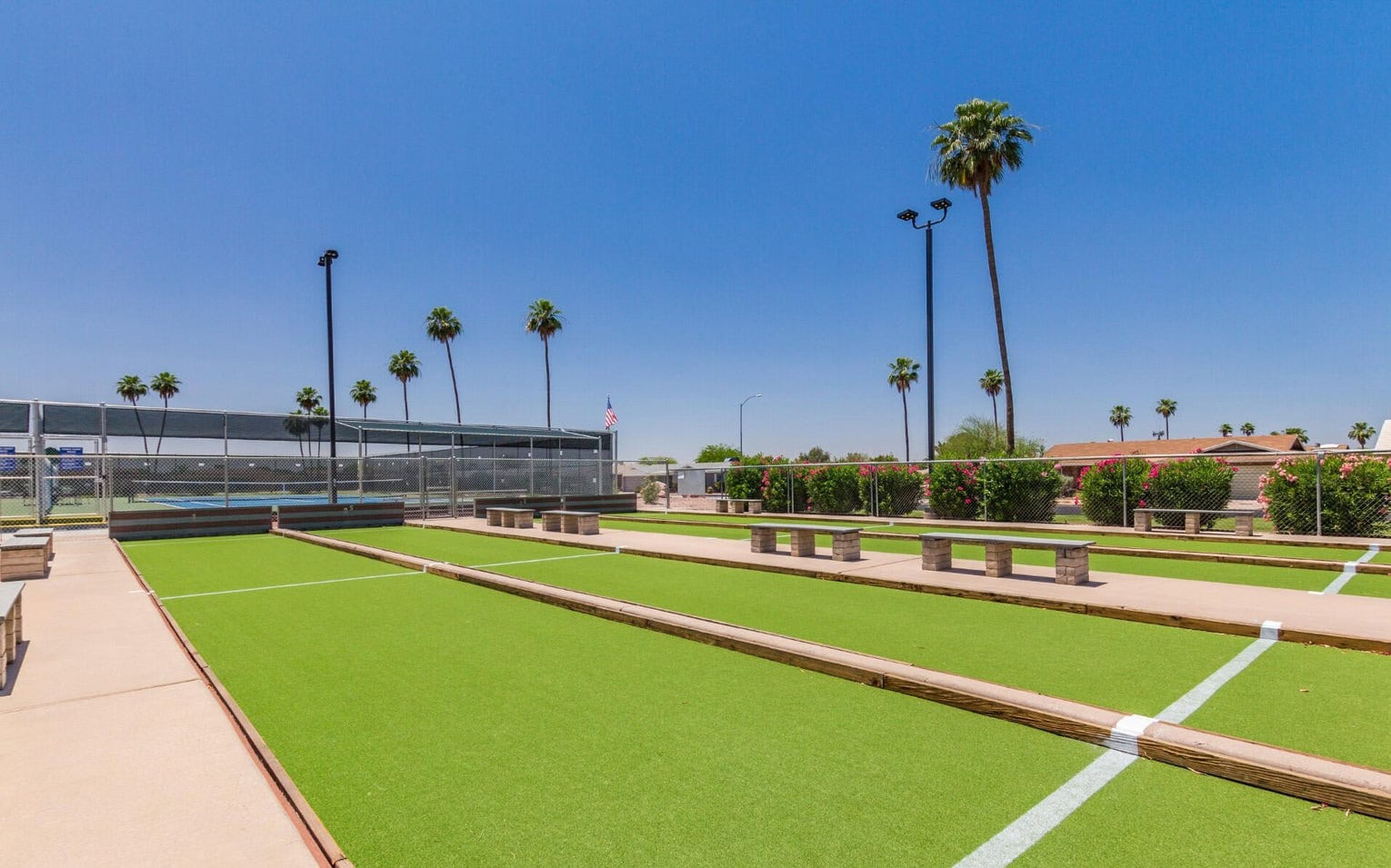 Outdoor bocce ball courts with artificial turf installations are lined with wooden borders. The area features benches for seating and is surrounded by palm trees under a clear blue Scottsdale sky. A net barrier stands in the background.