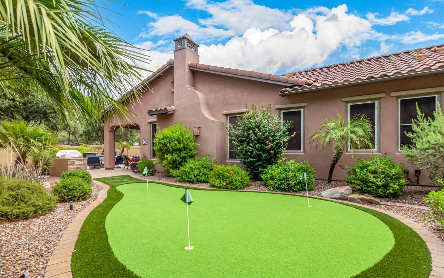 A suburban house with a red-tiled roof, nestled in Paradise Valley, AZ, is surrounded by lush greenery. In the foreground, a small putting green with golf flags features immaculate artificial grass, bordered by rocks and plants under a bright blue sky with fluffy clouds.