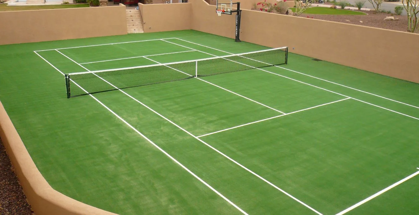 Nestled in the serene landscape of Scottsdale, this outdoor court features green artificial turf with crisp white lines for tennis and a basketball hoop at one end. Encircled by beige walls, it showcases the striking desert landscaping of Paradise Valley in the background.